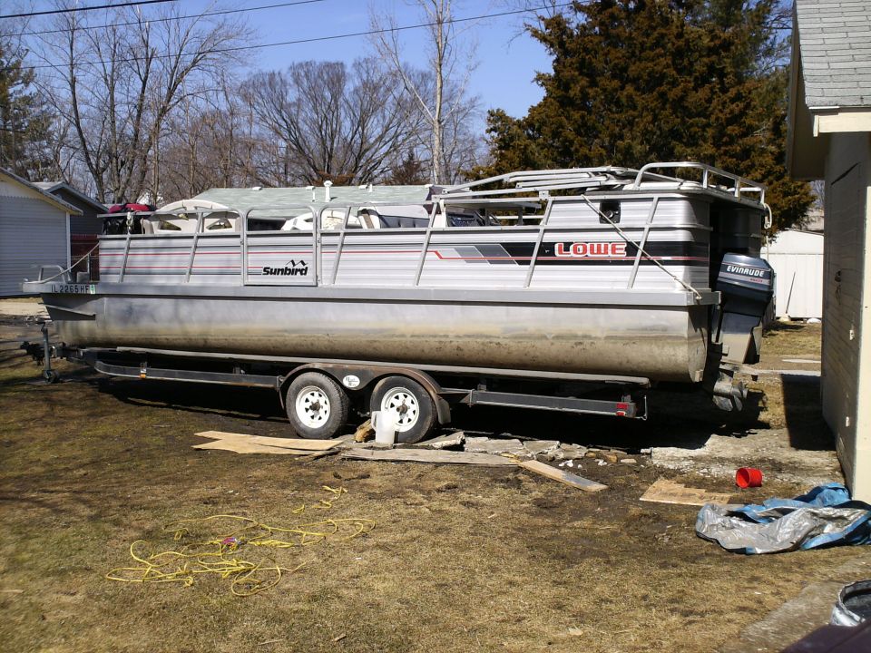 Pontoon boat with 'Sunbird' and 'Lowe' branding on a trailer, parked on a grassy area next to a house