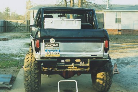 1978 Ford Bronco - Straight-on rear view of a lifted topless Ford Bronco parked on a residential driveway, showing a dented white tailgate with FORD lettering…