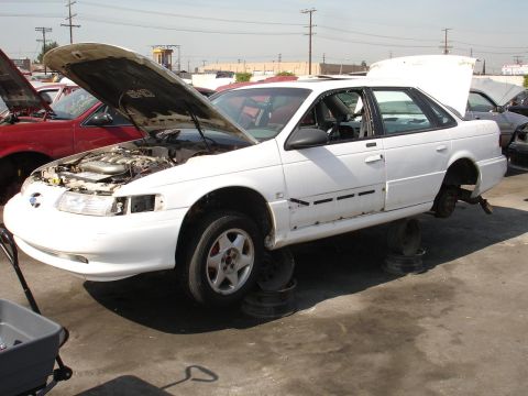 1993 Ford Taurus - White 1993 Ford Taurus sedan missing wheels, hood and trunk open, in a junkyard.