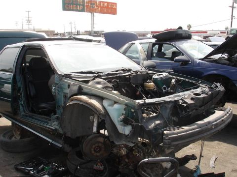 1993 Ford Taurus - A dismantled 1993 Ford Taurus coupe sits in a junkyard, its engine and front end removed.