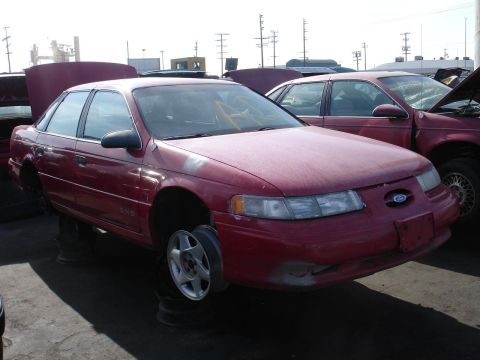 1993 Ford Taurus - A red sedan sits in a junkyard with its hood up and wheels removed.