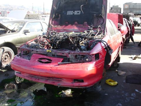 1993 Ford Taurus - A red 1993 Ford Taurus SHO sits in a junkyard with its hood open, revealing a dismantled engine and exposed wiring.