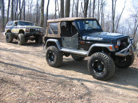 1992 Ford Mustang - Two modified off-road vehicles, a Jeep Wrangler and a Ford Bronco, are parked on a dirt path surrounded by trees.