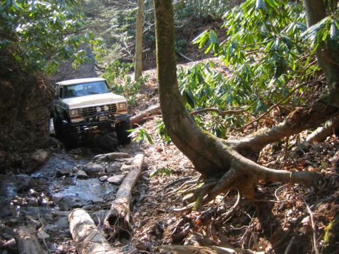 1992 Ford Mustang - A tan 1980s Ford Bronco navigates a challenging rocky stream bed surrounded by dense forest foliage.