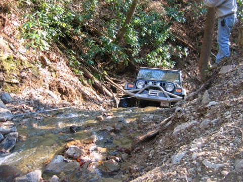 1992 Ford Mustang - A Jeep navigates a challenging off-road path, crossing a shallow stream with rocky banks.