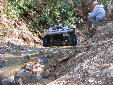 1992 Ford Mustang - A man watches an off-road vehicle navigate a challenging stream crossing on a wooded trail.