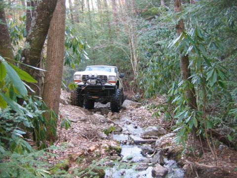 1992 Ford Mustang - Modified truck driving off-road through a shallow stream in a wooded area.