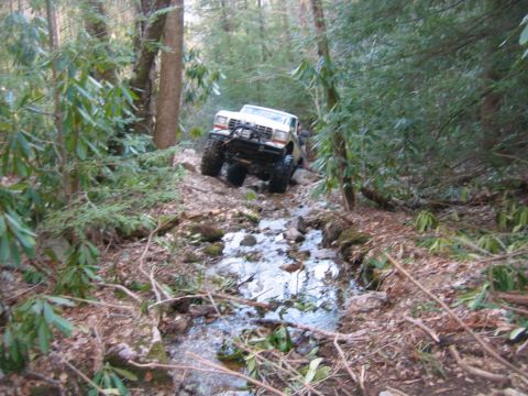 1992 Ford Mustang - This modified pickup truck navigates a challenging off-road path through a shallow stream in a forest.