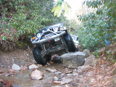 1992 Ford Mustang - An off-road Jeep navigates a rocky stream bed surrounded by dense foliage.