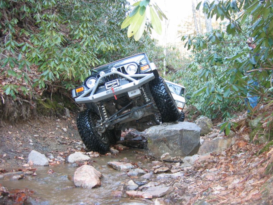 Jeep crawling over rocks in a stream, another white vehicle behind it