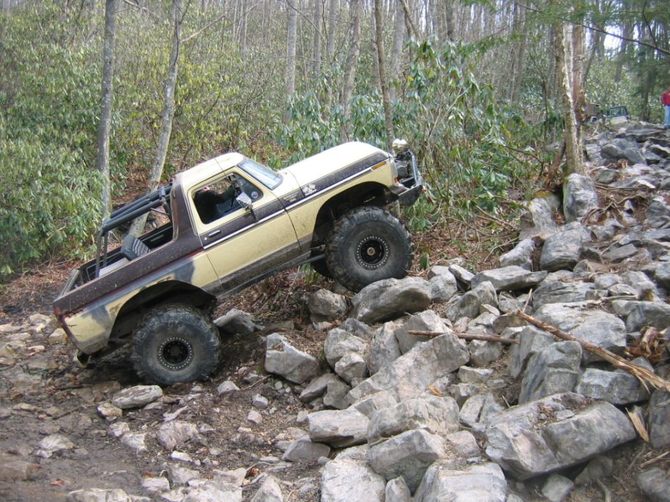 Tan and brown off-road vehicle with large tires climbing over rocks on a wooded trail