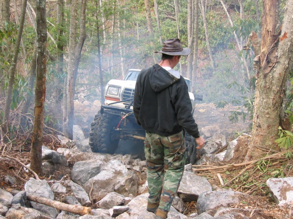 Man in hat and camouflage pants walks on rocks near a lifted off-road vehicle with large tires in a wooded area