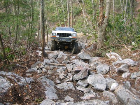 1992 Ford Mustang - A white off-road vehicle carefully maneuvers through a challenging rocky path surrounded by trees.