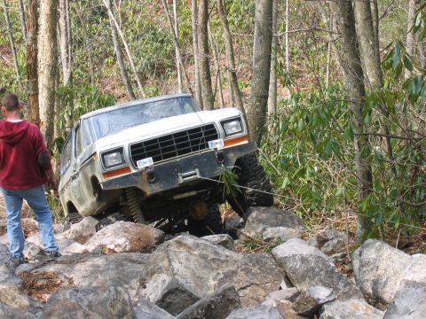 1992 Ford Mustang - A white Ford Bronco navigates a rocky trail in a wooded area, with a person observing the off-road challenge.