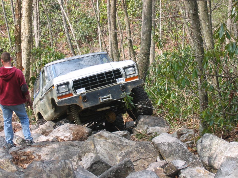 White Ford Bronco climbing over rocks on a wooded trail. Guy in red hoodie watches