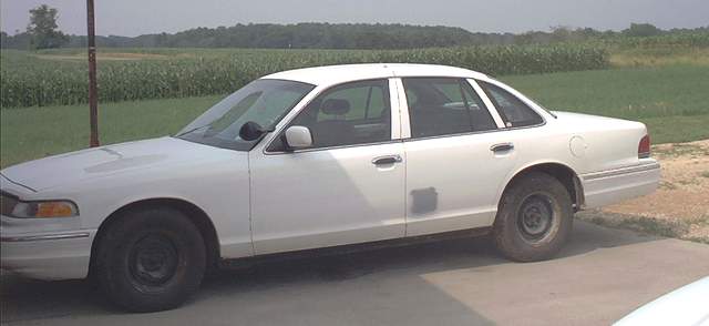 White Ford Crown Victoria sedan side view on gravel with cornfield behind