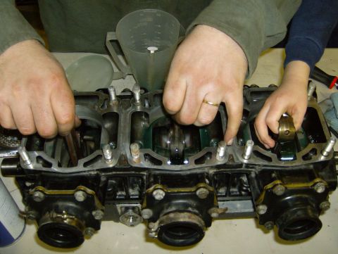 1982 Chevrolet C/K Pickup - Hands work on assembling an engine block, with a measuring cup and funnel nearby.