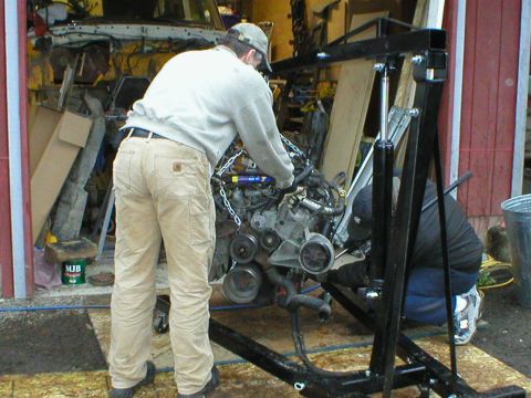1978 Ford Bronco - Two mechanics work on an engine suspended from an engine hoist in a garage setting.