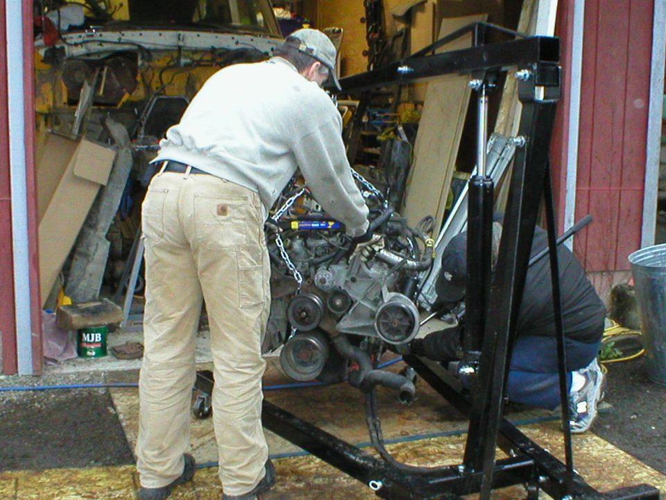 Man removing a V-engine on a black hydraulic engine hoist in a garage doorway.