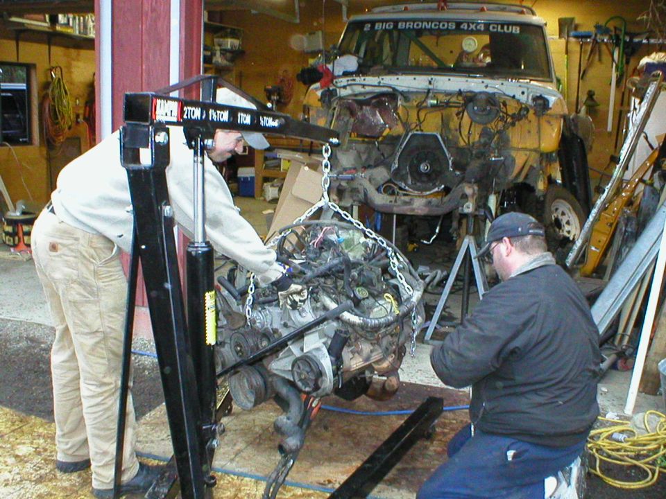 V8 engine hung on hoist chains with two men, stripped Bronco on jack stands behind in garage