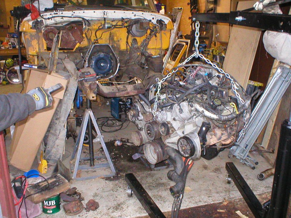 Engine suspended from hoist chain in garage beside stripped vehicle body