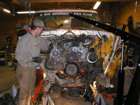 1978 Ford Bronco - A mechanic works on removing an engine from a white truck in a cluttered garage.