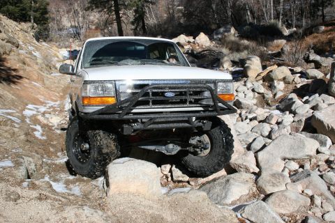 1996 Ford Bronco - White 1996 Ford Bronco navigates a rocky off-road trail with snow patches.
