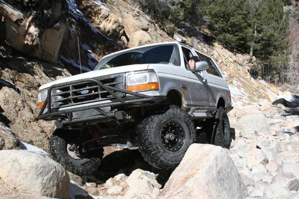 White and silver Ford Bronco with aftermarket bumper and large tires navigating rocky terrain
