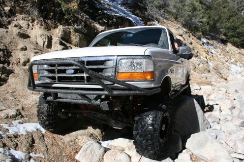 1996 Ford Bronco - A white 1996 Ford Bronco navigates a rocky, off-road trail with snow patches on the ground.