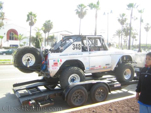 1990 Ford Bronco - white early Ford Bronco race truck marked 348 and Hermanos Cervantes Racing sits on a trailer as a boy stands beside it.