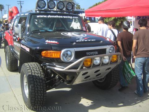 1990 Ford Bronco - black Toyota FJ Cruiser prerunner with roof-mounted lights and a tube bumper draws a crowd at an outdoor event.