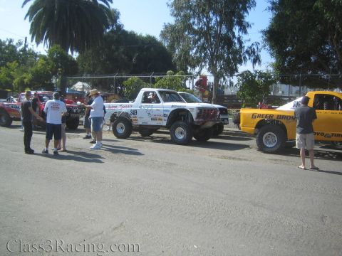 1990 Ford Bronco - white Ford race truck numbered 349 and a yellow truck numbered 806 sit on a dirt lot as people stand around them.