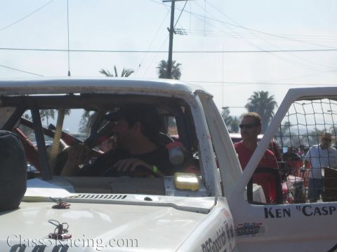 1990 Ford Bronco - driver sits inside the cab of a white off-road race truck while bystanders watch under a hazy sky with palm trees.