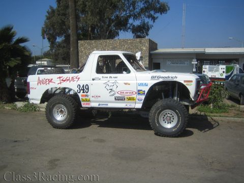 1990 Ford Bronco - Side profile of white Ford race truck numbered 349 lettered Anger Issues parked on pavement outside a stone building.