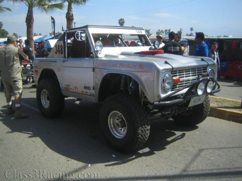 1990 Ford Bronco - silver early Ford Bronco race truck numbered 348 and lettered West Coast Broncos sits among a crowd at an outdoor event.