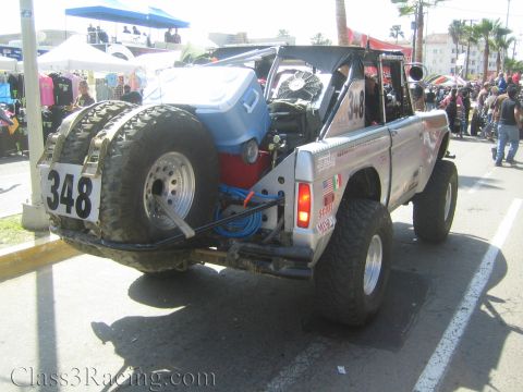 1990 Ford Bronco - Rear three-quarter view of silver Ford Bronco race truck 348 showing a rear-mounted spare tire and engine cooling fan.