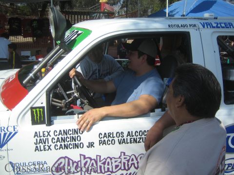 1990 Ford Bronco - Men sit in the cab of a white race truck covered in sponsor decals including driver names and a Monster logo at an event.