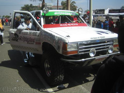 1990 Ford Bronco - white lifted Ford off-road race truck marked 'Chupakabras Racing' and 'Hecho en Mexico' sits in a crowded street paddock with a driver stan…