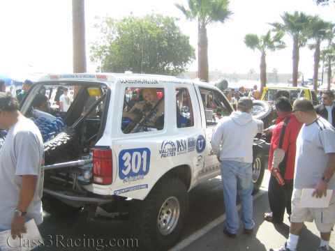 1990 Ford Bronco - rear of a white off-road race truck numbered 301 with caged interior and tall tires, surrounded by spectators on a sunny paved lot lined wi…