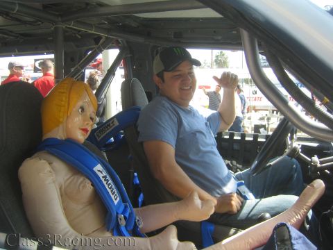 1990 Ford Bronco - grinning man in a Monster Energy cap sits buckled into a tube-frame race truck with an inflatable doll strapped into the passenger seat.