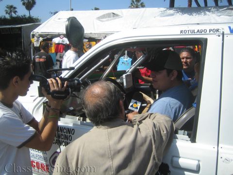 1990 Ford Bronco - cameraman and onlookers crowd around the open window of a white race truck as a man in a cap holds a microphone for an interview.