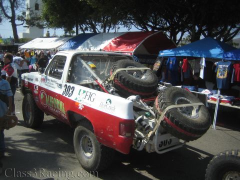 1990 Ford Bronco - rear of a red and white off-road race truck numbered 303 carries two spare tires on a tube rack, parked near vendor canopies and trees.