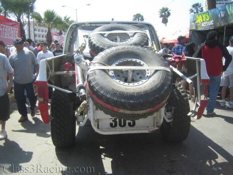 1990 Ford Bronco - Rear view of a white off-road race truck numbered 303 with mounted spare tires and a tube frame, surrounded by spectators on a street.