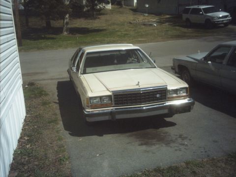 1985 Ford Crown Victoria LTD - A beige Ford Crown Victoria is parked in a driveway next to another car, with an SUV in the background.