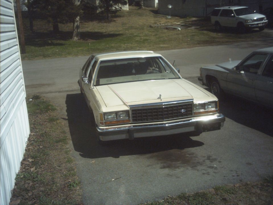 Cream-colored Ford Crown Victoria parked in driveway, another car and SUV visible