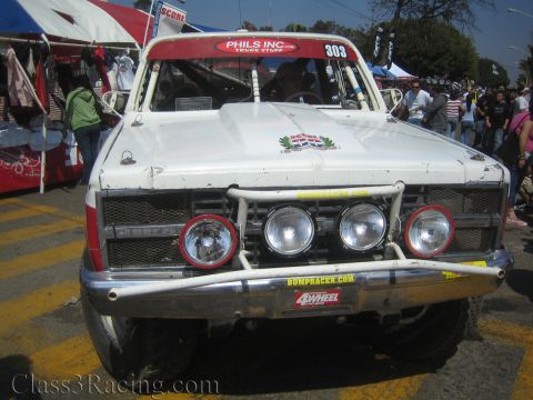 1990 Ford Bronco - Front of a weathered white 'Phils Inc' number 303 off-road race truck with four round driving lights and a tube bumper at a vendor market.