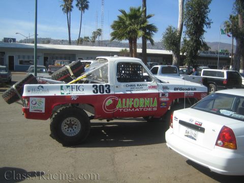 1990 Ford Bronco - Side view of a red and white 'California Tomatoes' number 303 off-road race truck with spare tires, parked next to a white sedan by buildin…