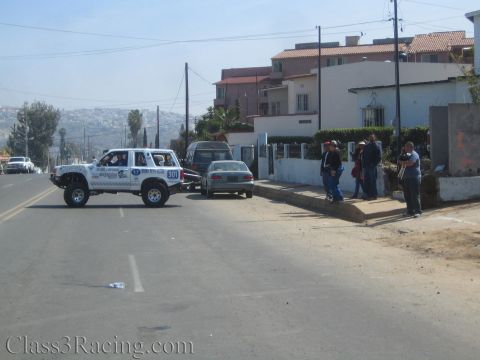 1990 Ford Bronco - white lifted race SUV crosses a hilltop residential street as bystanders watch from the sidewalk.