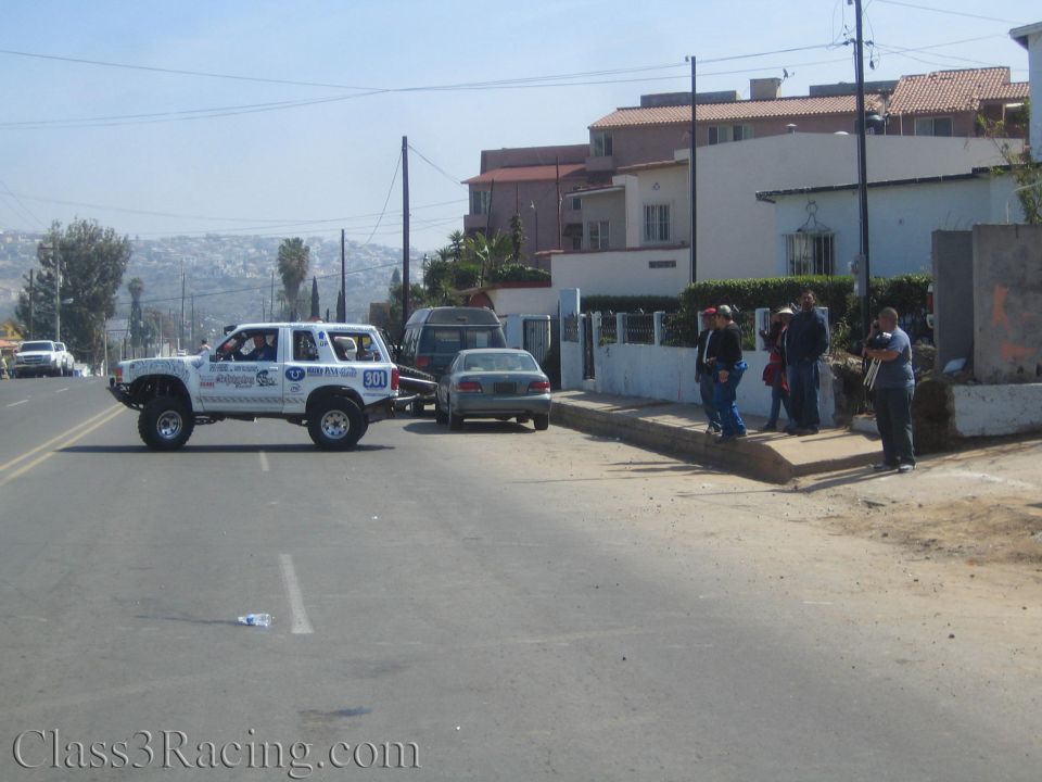White lifted SUV crossing a street near hillside houses with onlookers