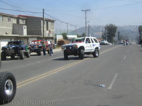 1990 Ford Bronco - white lifted SUV and several off-road trucks line up on a residential street with hills in the background.